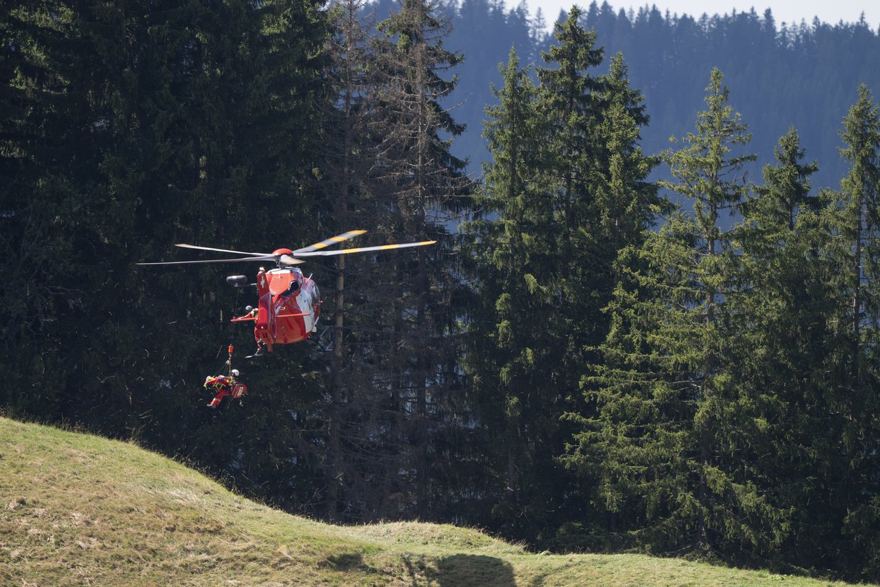 Hélicoptère de sauvetage rouge de la Rega effectuant une manœuvre de treuillage au-dessus d'une clairière boisée, avec une médecin suspendue au treuil.