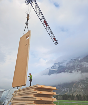 Bauarbeiter dirigiert ein grosses, mit einem Kran gehobenes Holzelement für die Rega-Basis Mollis vor einer Kulisse aus nebelverhangenen Bergen.