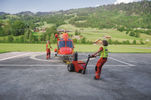 Zwei Rega-Crew-Mitglieder, darunter Jacqueline Zbaeren, manövrieren einen Rettungshelikopter mit einem Schlepper auf dem Helipad der Basis.