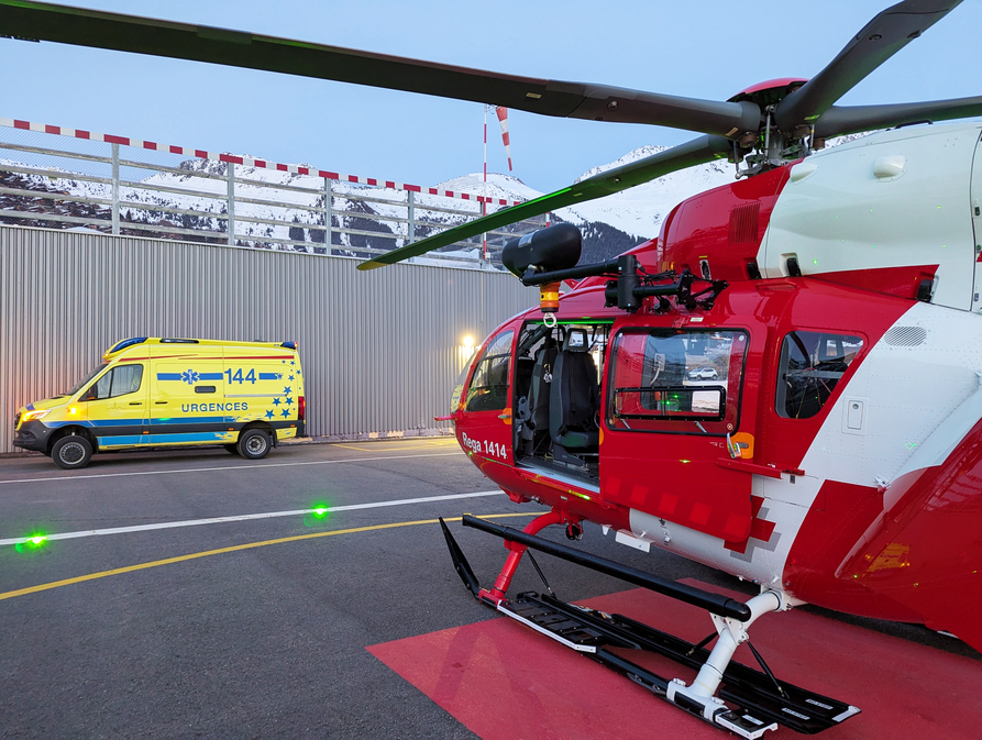 Un hélicoptère de la Rega se trouve sur l'héliport ; une ambulance est garée en face.