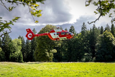 Zum Downloadformular für das Bild Rega-Helikopter schwebt tief über einer Wiese am Rand eines Waldes.