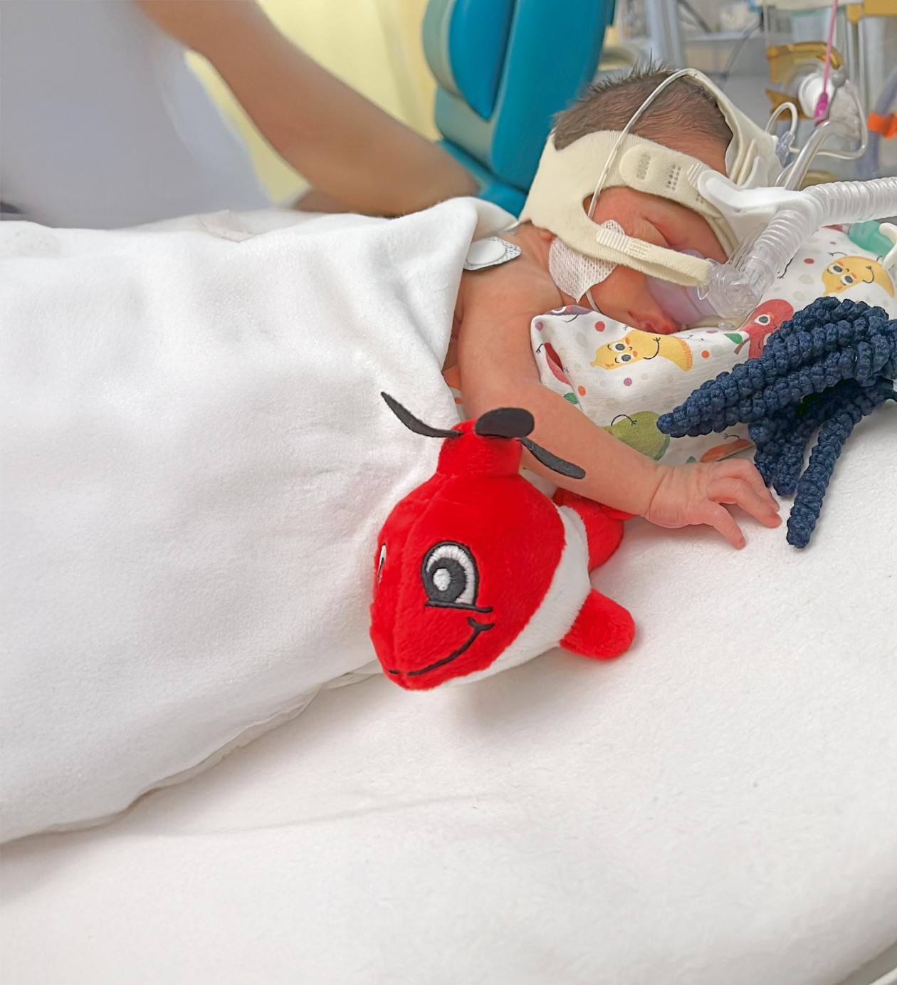 Un bébé est allongé dans un incubateur avec un appareil médical sur la tête, à côté d'une peluche rouge en forme d'hélicoptère.