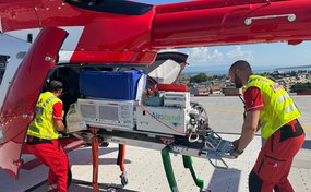 Deux collaborateurs de la Rega chargent un hélicoptère de sauvetage avec du matériel médical, dont un incubateur, pour une mission. La scène se déroule sur une héliplateforme.