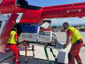 Deux collaborateurs de la Rega chargent un hélicoptère de sauvetage avec du matériel médical, dont un incubateur, pour une mission. La scène se déroule sur une héliplateforme.