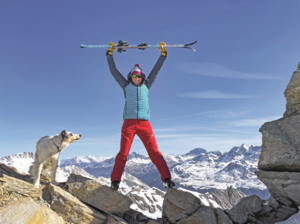 Rega-Rettungssanitäterin Jacqueline Zbaeren in ihrer Freizeit auf einem Gipfel in den Bergen, Skier triumphierend über den Kopf haltend, neben ihr ein Hund.