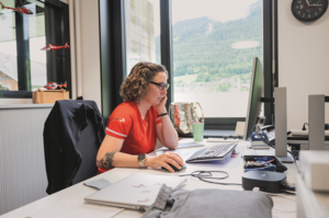 Rega-Rettungssanitäterin Jacqueline Zbaeren bei Büroarbeiten am Computer in der Basis, mit Blick auf die Schweizer Bergwelt.