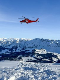 Ein roter Rettungshelikopter fliegt über eine verschneite Berglandschaft und zieht ein dünnes Seil nach unten. Im Hintergrund erstrecken sich weite Alpenpanoramen bei klarem Himmel.
