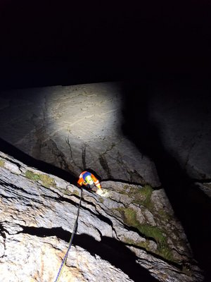 Un secouriste de la Rega descend en rappel la nuit, sur une paroi rocheuse et abrupte. Un projecteur éclaire la paroi, et le sauveteur porte un casque et une combinaison d'escalade bleue, orange et jaune.
