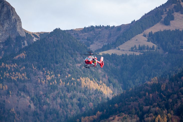 Hélicoptère rouge de la Rega en vol devant un vaste paysage montagneux avec des forêts aux couleurs automnales.