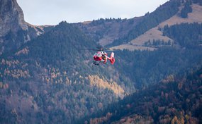 Hélicoptère rouge de la Rega en vol devant un vaste paysage montagneux avec des forêts aux couleurs automnales.