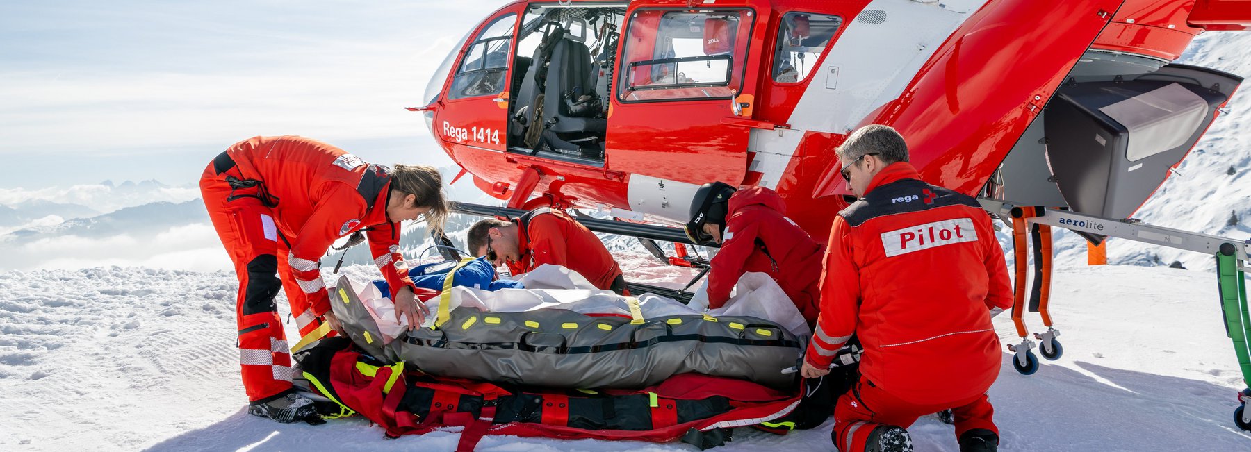 Eine Rega-Crew (Pilot und medizinische Crew) versorgt einen Patienten auf einer Schneeunterlage neben einem roten und weissen Airbus H145 Helikopter in einer weissen, sonnigen Berglandschaft.