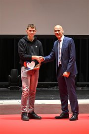 Due uomini si stringono la mano su un palco. L'uomo a sinistra, Lukas Piffero, riceve un premio a forma di cuore dall'uomo a destra, Gabriele Casso, che indossa un abito.