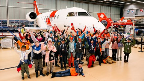 Eine grosse Gruppe von Kindern und Erwachsenen jubelt in einem Hangar. Im Hintergrund stehen ein Ambulanzjet und zwei Helikopter der Rega.