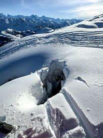 Eine tiefe Spalte im Schnee öffnet sich inmitten einer unberührten Winterlandschaft. Die umliegenden Berge sind in helles Sonnenlicht getaucht.