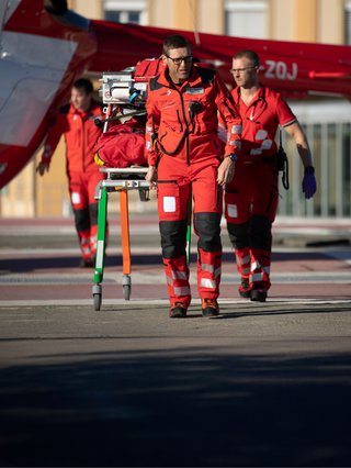 Two Rega employees in red uniforms transport a stretcher with medical equipment, with a helicopter in the background.