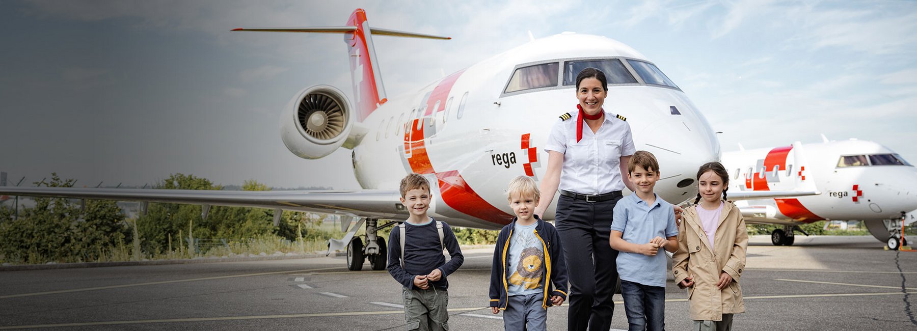 Eine Rega-Pilotin steht lächelnd mit vier Kindern vor einem Rega-Ambulanzjet.