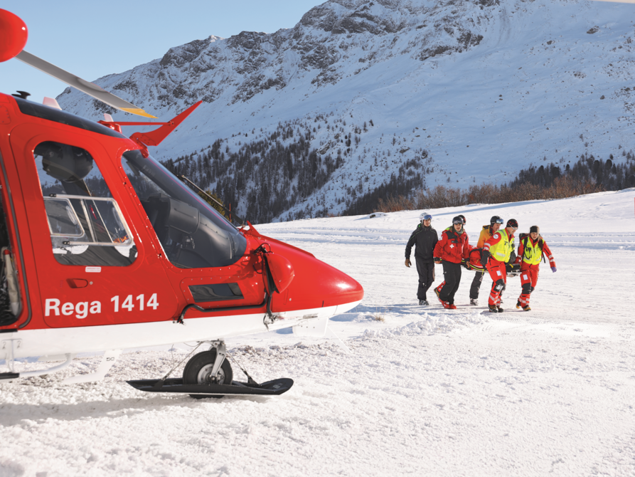 A Rega crew transports a person on a stretcher across a snow-covered mountain landscape towards the waiting rescue helicopter.
