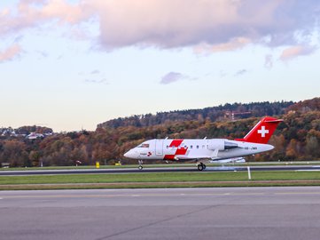 Rega-Ambulanzjet landet auf einer Start- und Landebahn vor bewaldeten Hügeln in herbstlicher Landschaft.
