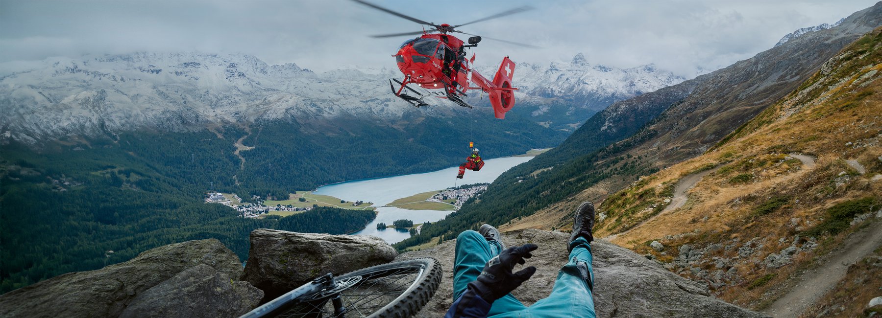 Ein Rega-Helikopter seilt eine Person mit einer Trage über einem See und einer Berglandschaft ab, aus der Perspektive einer Person, die auf einem Felsen liegt und ein Fahrrad neben sich hat.