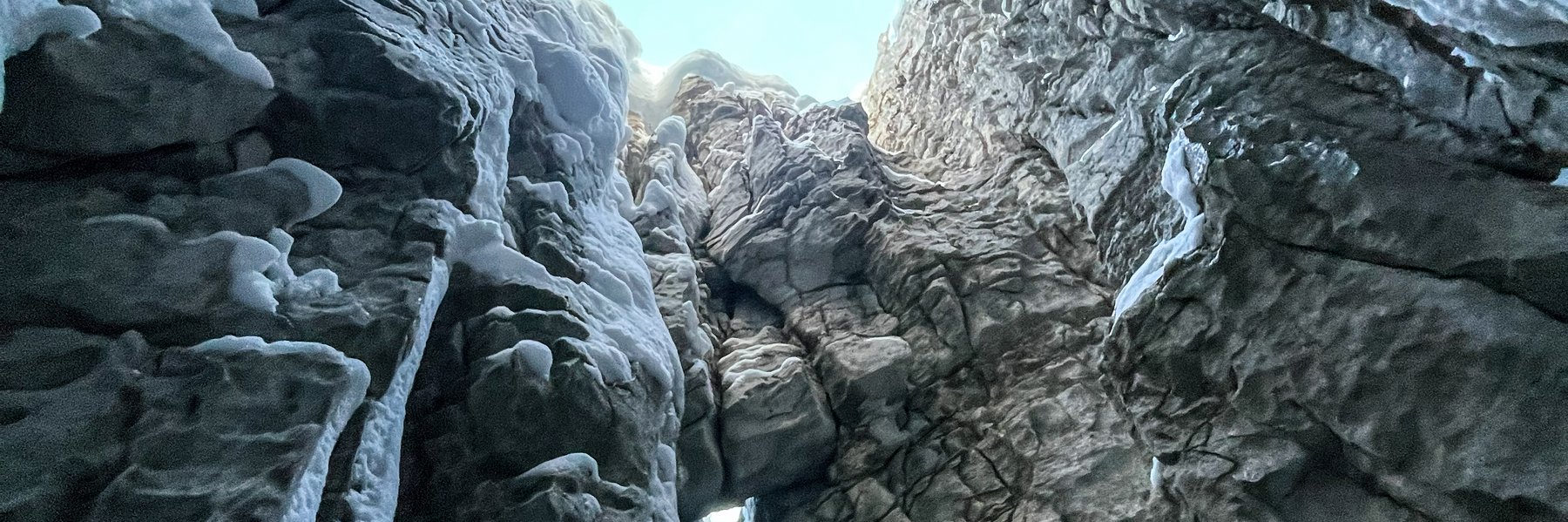 Blick aus einer tiefen, engen Felsspalte nach oben zum Tageslicht, mit vereisten Felswänden rundherum. Das Eis hängt in langen Strukturen von den Felsen herab.