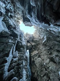 Blick aus einer tiefen, engen Felsspalte nach oben zum Tageslicht, mit vereisten Felswänden rundherum. Das Eis hängt in langen Strukturen von den Felsen herab.