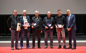 Sei uomini sono in piedi su un palco con il pavimento rosso. Quattro di loro tengono in mano dei premi a forma di cuore, probabilmente un riconoscimento per primi soccorritori.