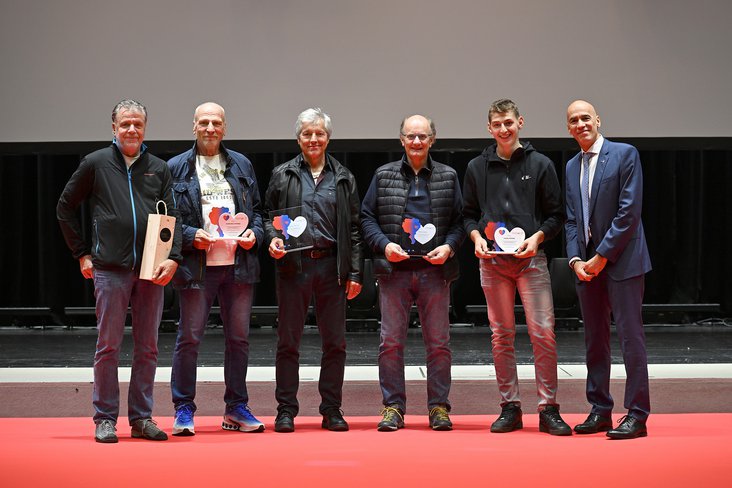 Sei uomini sono in piedi su un palco con il pavimento rosso. Quattro di loro tengono in mano dei premi a forma di cuore, probabilmente un riconoscimento per primi soccorritori.