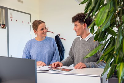 Ein Mediamatiker-Lehrling sitzt an einem Tisch im Gespräch mit einer jungen Mitarbeiterin. Vor ihnen liegt ein aufgeschlagenes Rega-Magazin zur Besprechung.