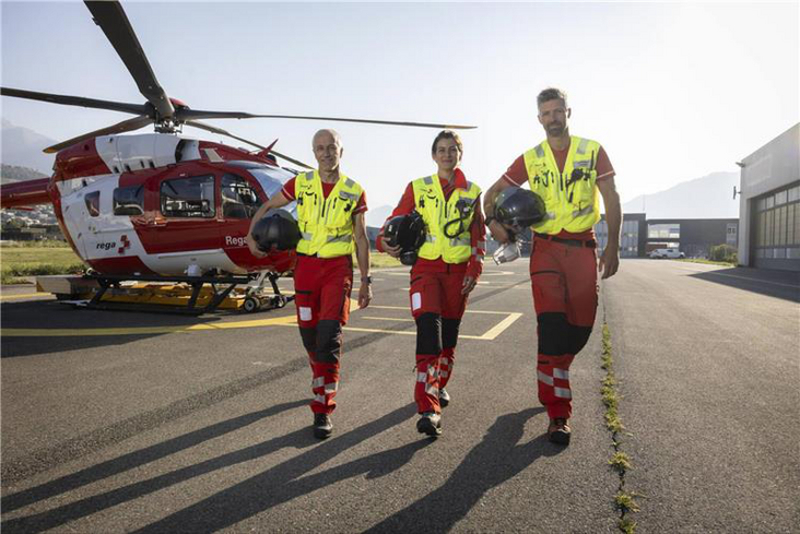 Drei Rega-Crewmitglieder gehen in ihren roten Einsatzanzügen und gelben Warnwesten auf dem Helikopterlandeplatz. Im Hintergrund steht ein Rega-Helikopter vom Typ Airbus H145.