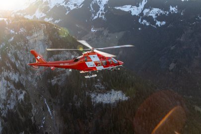 A Rega rescue helicopter flying past a steep cliff face in the Alps.