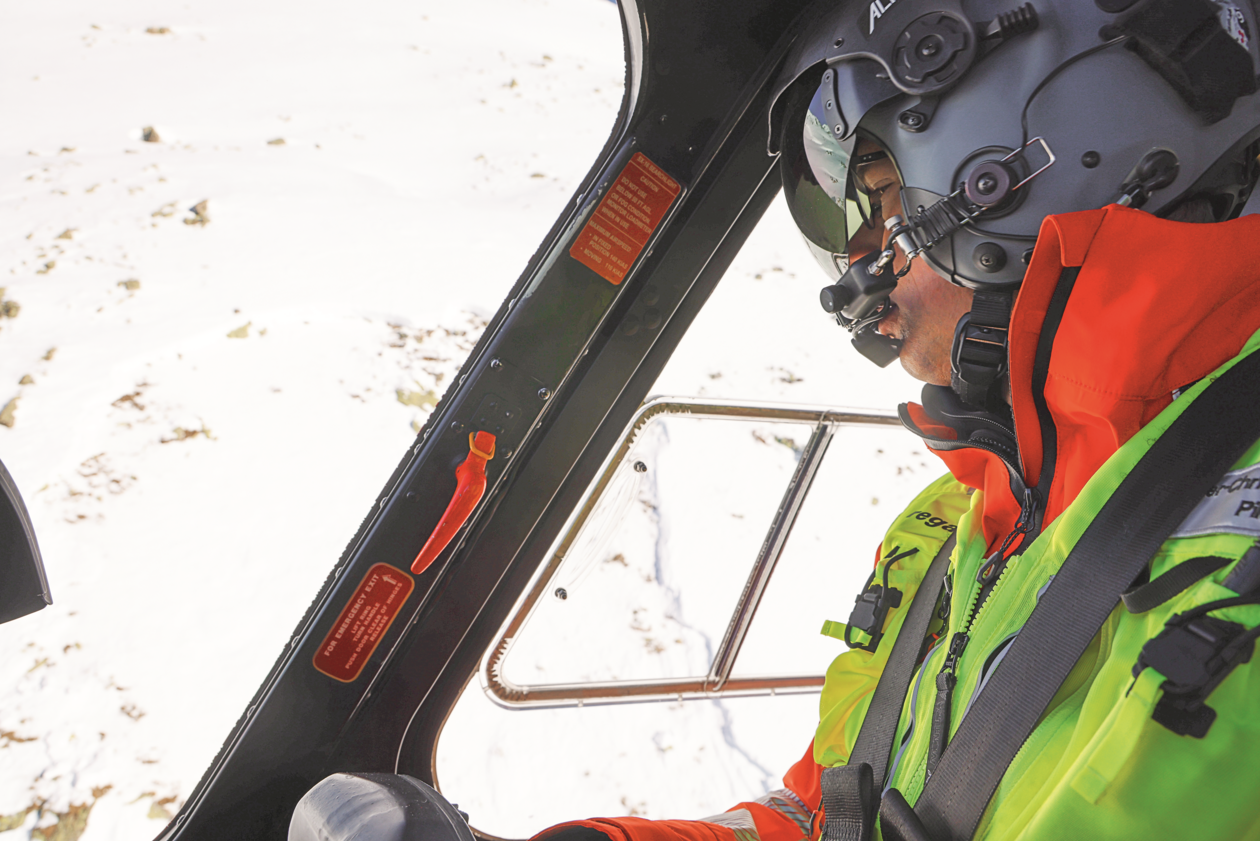 Close-up of a Rega pilot wearing a helmet and goggles inside the cockpit during a mission flight over a snowy mountain landscape.