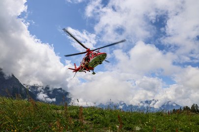 Un hélicoptère de sauvetage AgustaWestland Da Vinci de la Rega est en vol stationnaire à basse altitude au-dessus d'une prairie de montagne. La photo est prise d'une perspective basse, avec de vastes nuages et des montagnes en arrière-plan.
