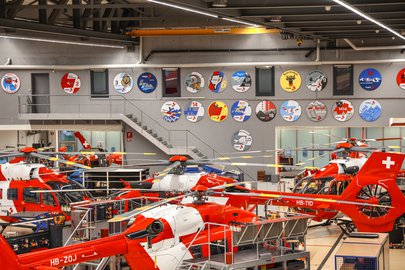 Plusieurs hélicoptères de la Rega en maintenance dans le hangar du Centre Rega ; les logos des bases sont affichés au mur.