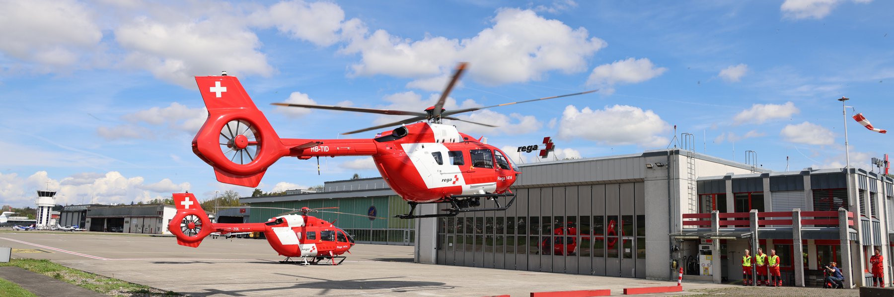 Elicottero H145 D3 Rega in volo o poco prima dell'atterraggio/decollo, con l'hangar Rega sullo sfondo e cielo azzurro con nuvole.