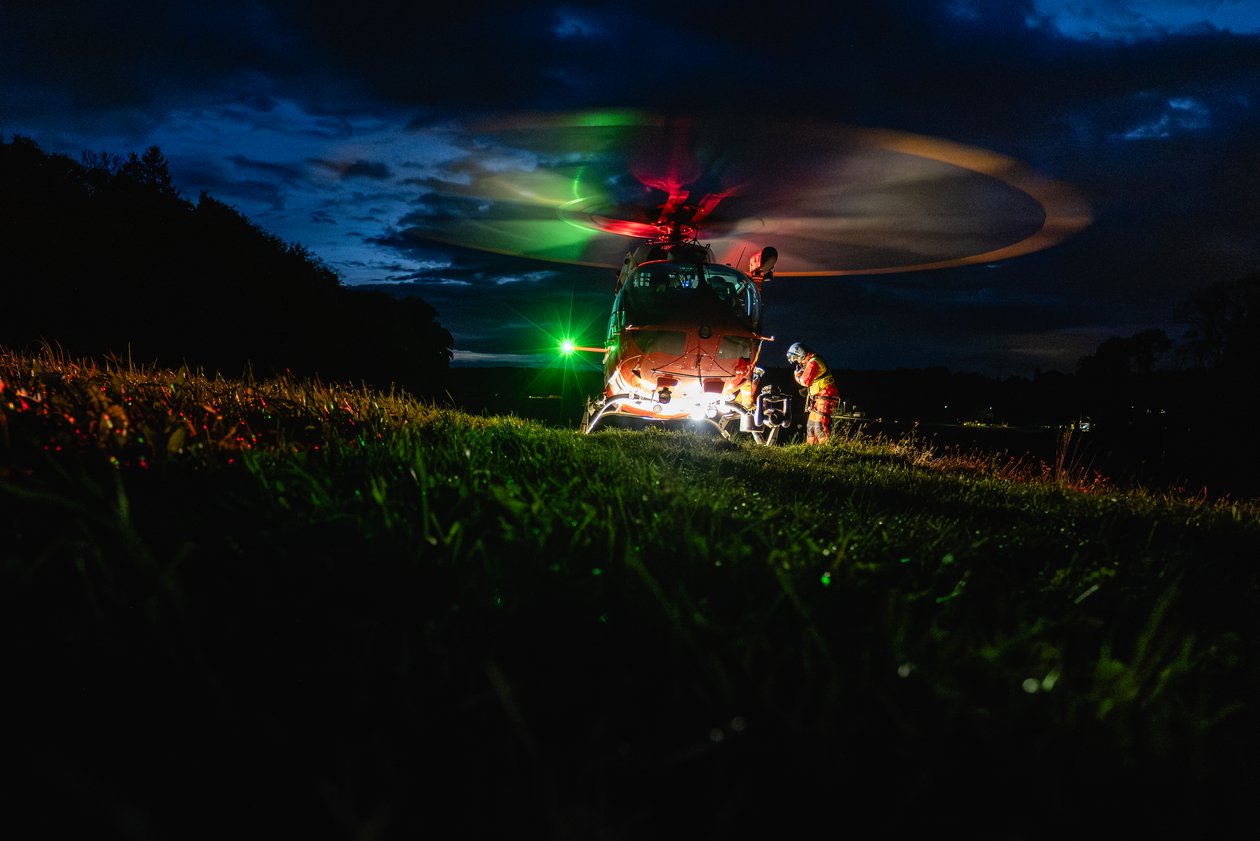 Hélicoptère de la Rega atterrissant de nuit sur une prairie sombre et herbeuse. Les lumières de l'hélicoptère et du sauveteur sont clairement visibles.