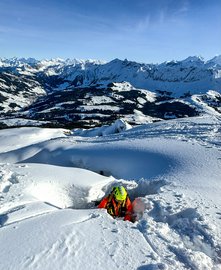 Eine Rettungsperson in orangefarbener Ausrüstung und gelbem Helm steigt aus einer Schneespalte hervor. Hinter ihr liegen weite Berge und ein verschneites Tal.