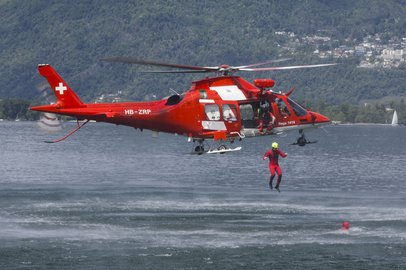 Un elicottero rosso della Rega (HB-ZRP) sorvola a bassa quota l'acqua, mentre un soccorritore viene calato in acqua. Sullo sfondo si vedono montagne boscose e costruzioni sulla riva.