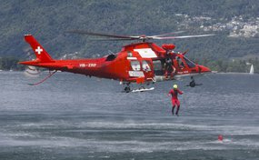 Un elicottero rosso della Rega (HB-ZRP) sorvola a bassa quota l'acqua, mentre un soccorritore viene calato in acqua. Sullo sfondo si vedono montagne boscose e costruzioni sulla riva.