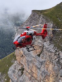 Un hélicoptère de sauvetage de la Rega vole près d'une paroi rocheuse abrupte dans les montagnes. Il a la livrée rouge et blanche typique de la Rega. Les membres de l'équipage sont visibles sur le côté gauche de l'image.