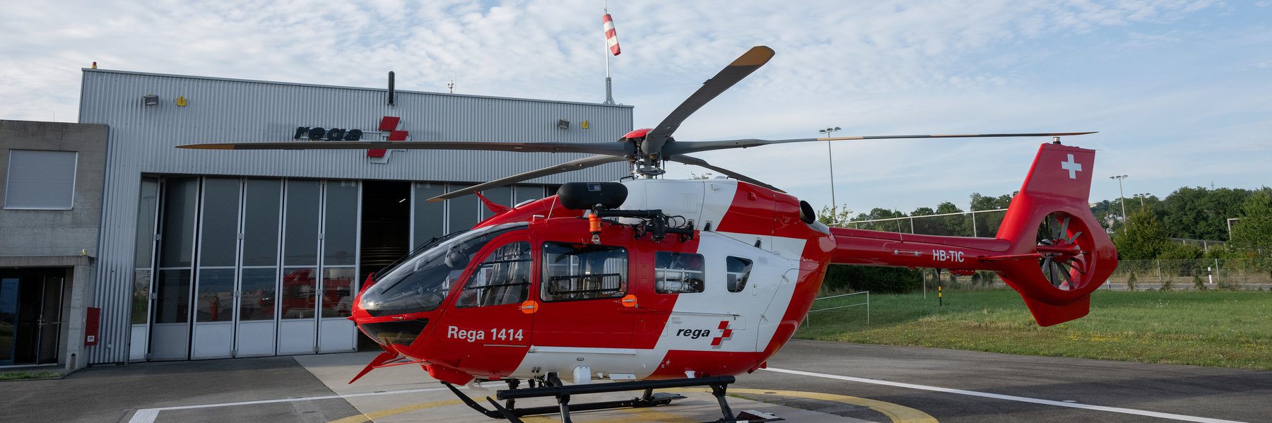 Rega H145 D3 helicopter in front of a hangar, ready for deployment under a cloudy sky. The Rega logo is visible on the hangar.