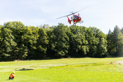 Un hélicoptère Rega H145 vole à basse altitude au-dessus d'une prairie. Un secouriste agenouillé dans l'herbe regarde l'hélicoptère. Un groupe d'arbres est visible en arrière-plan.