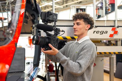 Ein Mediamatiker-Lehrling der Rega steht mit einer professionellen Videokamera in einem Hangar. Links ist ein Rega-Helikopter angeschnitten, im Hintergrund das Rega-Logo.