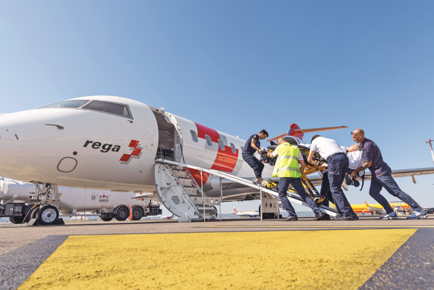Mehrere Rega-Mitarbeitende verladen einen Patienten auf einer Trage über die Treppe in den Ambulanzjet der Rega, der auf dem Rollfeld steht.