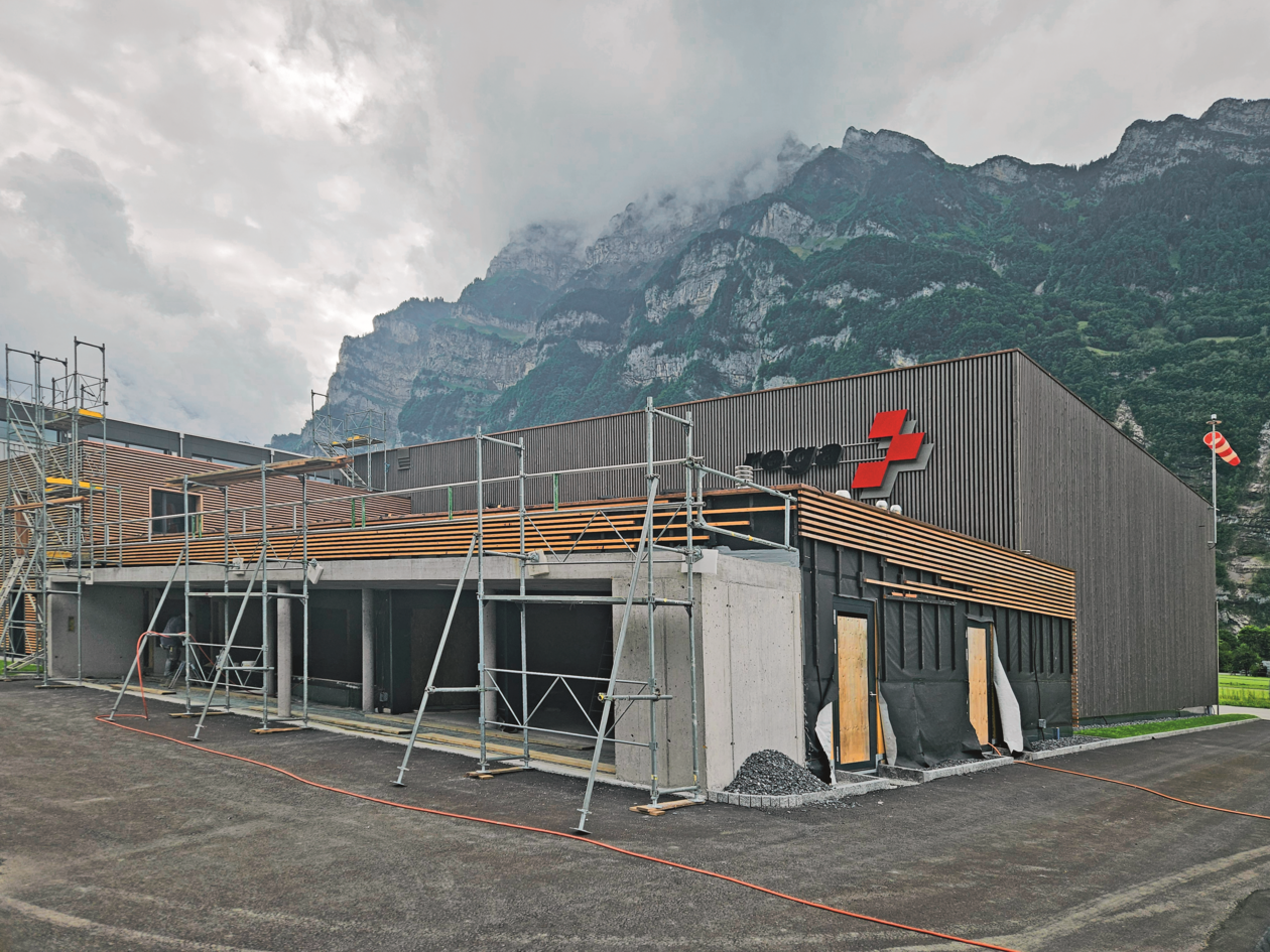 Aussensicht der Rega-Basis Mollis während der Bauphase, mit Gerüst vor der modern gestalteten Holzfassade und alpiner Kulisse.