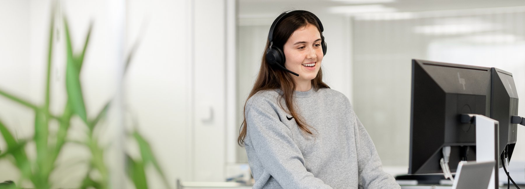 A smiling young woman wearing a headset is sitting at a desk and working on a computer. She represents the mission controllers at the operations center.