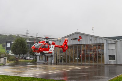 Ein rot-weisser Rega-Rettungshubschrauber hebt vor einem Hangar mit grossen Fenstern ab. Im Hintergrund sind weitere Helikopter und Ausrüstung sichtbar. Die nassen Oberflächen und der graue Himmel deuten auf einen regnerischen Tag hin.