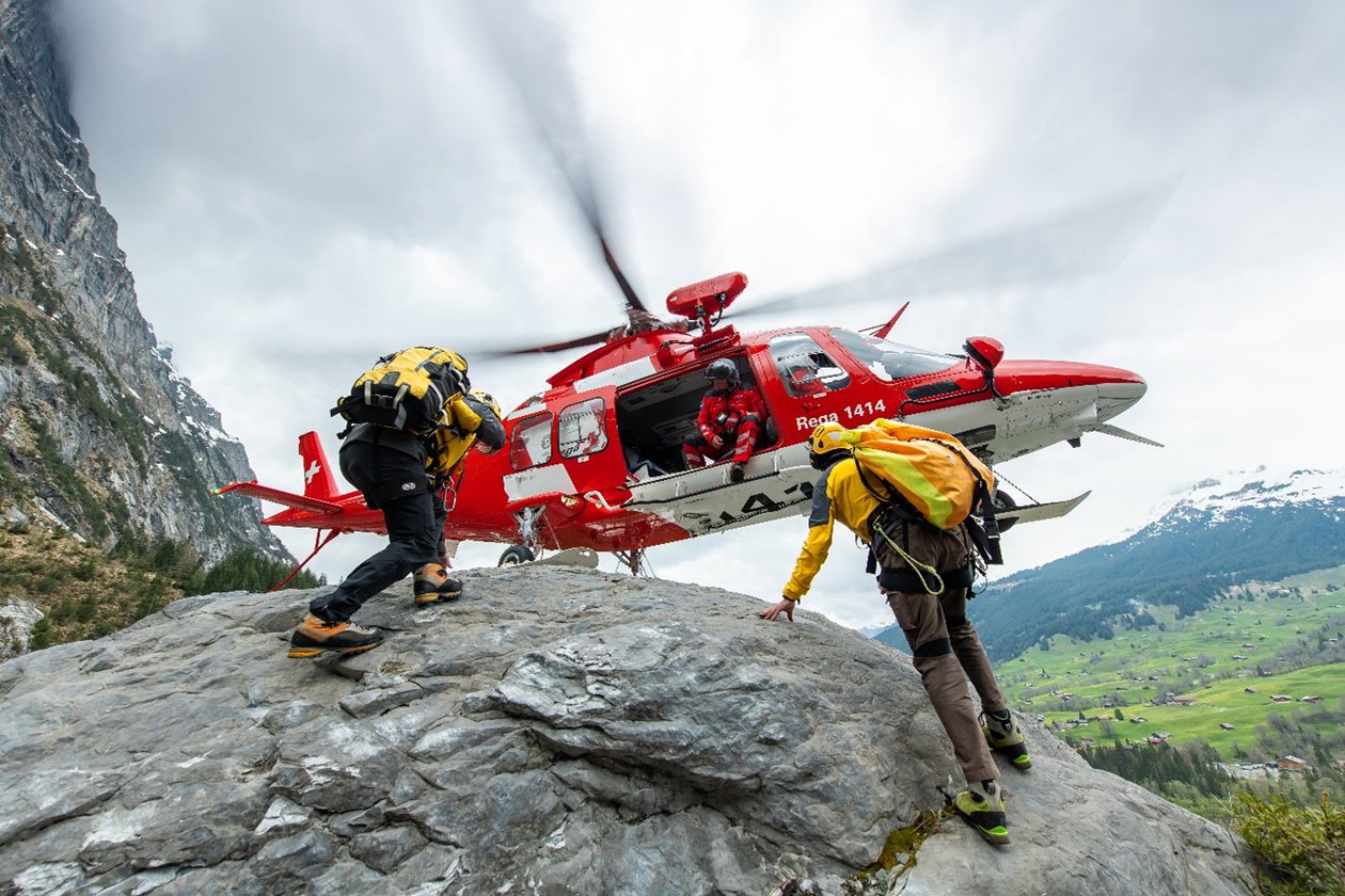 Ein Rettungshelikopter im Landeanflug vor dem zwei Bergretter des Schweizer Alpen-Club SAC knien 
