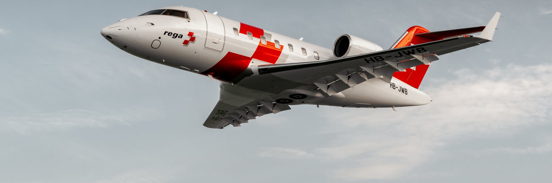 Ein weisser Rega-Ambulanzjet vom Typ Bombardier Challenger 650 mit rotem Kreuz im Flug vor bewölktem Himmel.