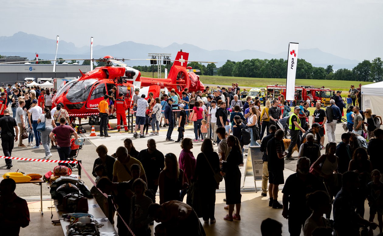 Eine grosse Menschenmenge versammelt sich auf einem Flugfeld um einen roten Rega-Rettungshelikopter bei einer öffentlichen Veranstaltung, gesehen aus einem Hangar.