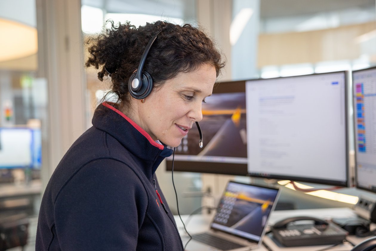 Une médecin-conseil de la Rega aux cheveux bouclés est assise dans la centrale d'opérations devant ses moniteurs. Elle porte un casque et sourit, prenant des appels et travaillant avec la souris sur l'un de ses écrans.
